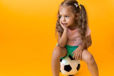 Klasik siyah-beyaz futbol topuyla gülümseyen sevimli anaokulu kızı sarı arka plandaki kameraya bakıyor. Çocuk tişört ve şortla futbol oynuyor. Spor, şampiyonluk, takım konsepti.