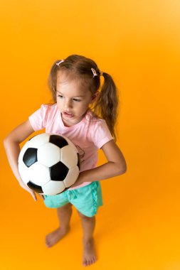 Klasik siyah-beyaz futbol topuyla gülümseyen sevimli anaokulu kızı sarı arka plandaki kameraya bakıyor. Çocuk tişört ve şortla futbol oynuyor. Spor, şampiyonluk, takım konsepti.