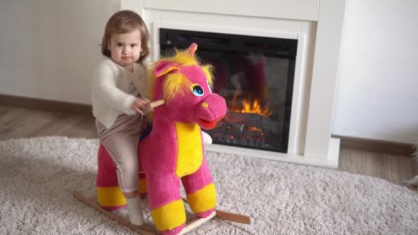 Familie jeugd. Happy kleine peuter kind meisje warme trui winter kleding spelen uitkleden op zacht paard in de kamer in de buurt van open haard. Leuke speelse authentieke Kid speelt thuis alleen swing op Toy — Stockvideo