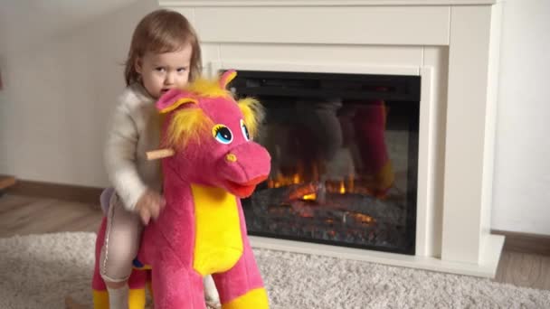 Familie jeugd. Happy kleine peuter kind meisje warme trui winter kleding spelen uitkleden op zacht paard in de kamer in de buurt van open haard. Leuke speelse authentieke Kid speelt thuis alleen swing op Toy — Stockvideo