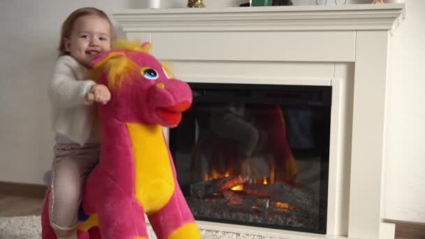 Family childhood. Happy little toddler child girl warm sweater winter clothes playing ridding on soft horse in room indoors near fireplace. Cute playful authentic Kid plays at home alone swing on Toy — Stock Video