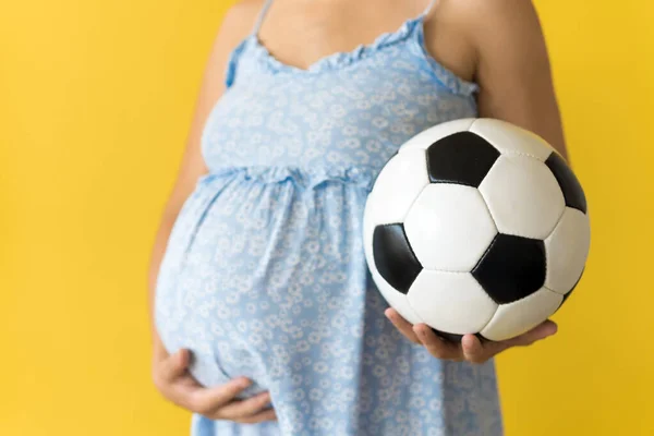 Motherhood, femininity, football, sport, dairy, hot summer. croped unrecognizable pregnant young pretty woman in floral blue dress holds soccer white and black ball rubs tummy on yellow background Royalty Free Stock Images