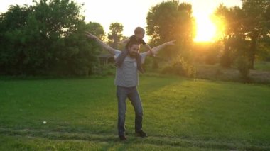 Yaz, tatil, doğa, mutlu aile, çocukluk, babalık, baba günü - küçük bir anaokulu çocuğuyla çocuk çocuk kanat çırpmaca oynuyor çocuklar günbatımında parktaki çayırda omuzlarına oturup eğleniyor.
