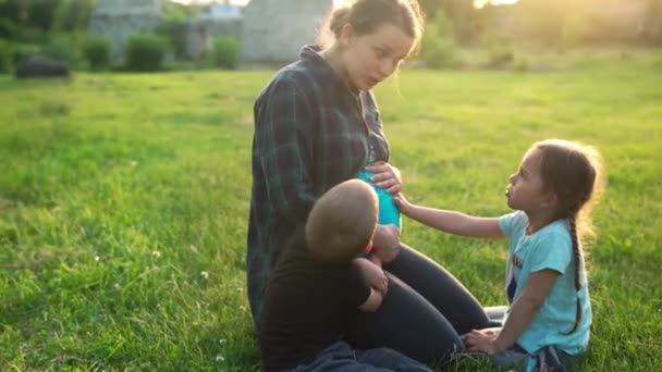 Літо, природа, щаслива сім'я, вагітність - молода вагітна мати з двома маленькими дітьми-малюками сидить на траві в парку на заході сонця. Діти погладжують маму поцілунок живота розважайтеся, смійтеся проводити час разом — стокове відео