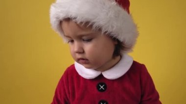 Şirin, mutlu, tombul, bebek bebek Noel Baba kostümlü, sarı arka plandaki kameraya bakıyor. Çocuk Noel Sahnesi Oynuyor Doğum gününü kutluyor. Çocuklar yeni yılı eğlenerek geçiriyorlar. Çocukluk