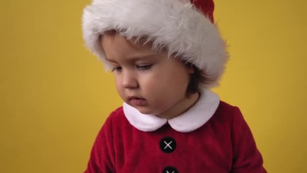 Симпатичная веселая девочка в костюме Санта-Клауса, смотрящая на камеру на желтом фоне. Ребенок играет в рождественскую сцену празднования дня рождения. Детям весело проводить новогоднее время. Детство — стоковое видео