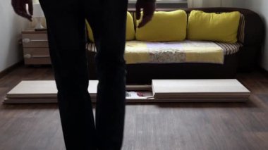 Young Black Bearded Long-haired Man Lays Out Parts Of Bookcase On Floor For Assembly Rack Furniture At Home. Collecting Wooden Boards On Living Room House. Moving At New Place. Guy Making Renovation.