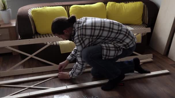 Young Black Bearded Long-haired Man Lays Out Parts Of Bookcase On Floor For Assembly Rack Furniture At Home. Collecting Wooden Boards On Living Room House. Moving At New Place. Guy Making Renovation. — стоковое видео