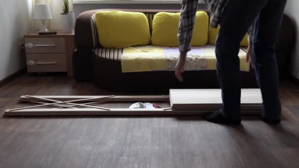 Young Black Bearded Long-haired Man Lays Out Parts Of Bookcase On Floor For Assembly Rack Furniture At Home. Collecting Wooden Boards On Living Room House. Moving At New Place. Guy Making Renovation. — стокове відео