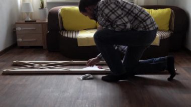 Young Black Bearded Long-haired Man Lays Out Parts Of Bookcase On Floor For Assembly Rack Furniture At Home. Collecting Wooden Boards On Living Room House. Moving At New Place. Guy Making Renovation.