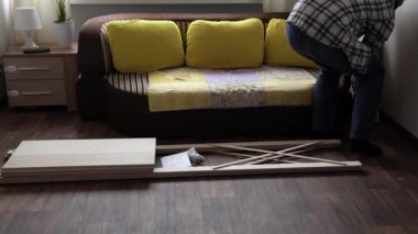 Young Black Bearded Long-haired Man Lays Out Parts Of Bookcase On Floor For Assembly Rack Furniture At Home. Collecting Wooden Boards On Living Room House. Moving At New Place. Guy Making Renovation.