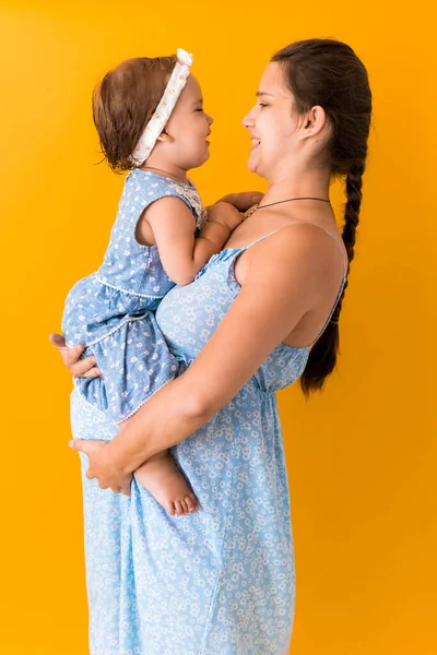 Материнство, жаркое лето - портрет молодой беременной счастливой улыбающейся матери женщины в синем платье, держа маленькую дочурку малыша, брата, босых ног в руках, глядя в глаза поцелуй на желтом фоне — стоковое фото