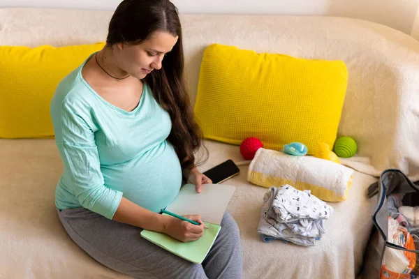 Wanita hamil imut menulis daftar kemasan untuk rumah sakit bersalin dengan tas Prepares Notebook. (Inggris) Young Ledy In Pregnancy Have Fun Spend Time On Sofa At Home. Keibuan, Kedokteran Konsep Kesehatan dan Perawatan Stok Gambar