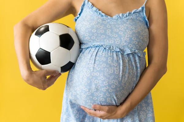 Motherhood, femininity, football, sport, dairy, hot summer. croped unrecognizable pregnant young pretty woman in floral blue dress holds soccer white and black ball rubs tummy on yellow background Royalty Free Stock Photos