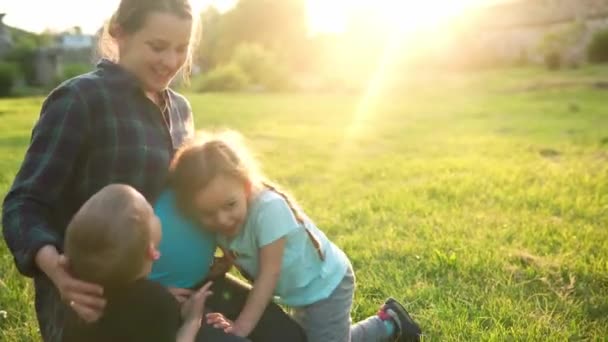 Léto, příroda, šťastná rodina, těhotenství - mladá těhotná matka se dvěma malými batolaty sedí při západu slunce v parku na trávě. Děti hladí maminku břicho polibek bavte se, smích trávit čas spolu — Stock video