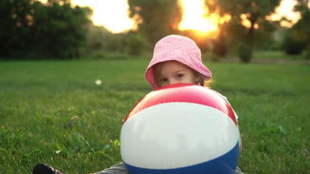 Verano, vacaciones, naturaleza, infancia feliz un niño pequeño niño preescolar sonriendo niña en sombrero rosa sentarse posando mirando el juego de la cámara con gran inflable en el parque en el prado al atardecer aire fresco — Vídeos de Stock