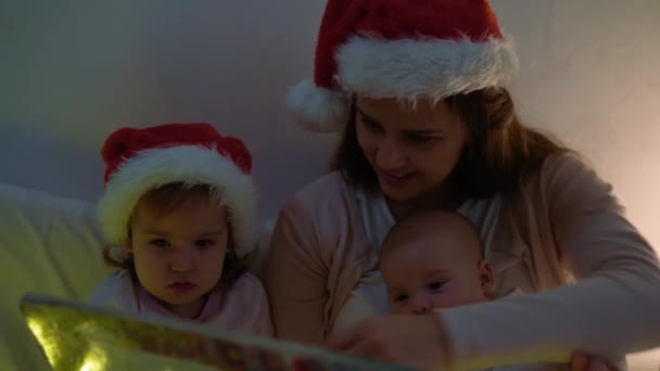 Szczęśliwa matka rodziny w czerwonym kapeluszu Mikołaja dwoje dzieci rodzeństwo małe rodzeństwo Boże Narodzenie noc w łóżku piżamy zabawy czytania bajki w ciemnym pokoju ciemności. Macierzyństwo, wakacje, dom zimowy, miłość — Wideo stockowe