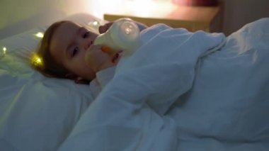 Genç anne yatak odası gecesinde küçük bebeğe biberonundan süt içiriyor. Kadın yeni doğmuş şirin bebeğe yumuşak ve sıcak bir şekilde dokunuyor. Anne, çocuğuna iyi bak.