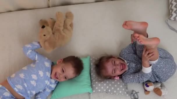 Dos niños de preescolar sonrientes en pijama jugando con el oso de peluche en la cama. Los hermanos Little Twins Boy y Girl se divierten. Niños felices en cuarentena en casa. Amistad, Familia, Concepto de Educación — Vídeos de Stock