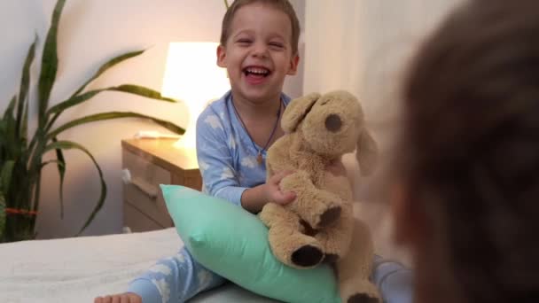 Deux enfants souriants d'âge préscolaire en pyjama jouant avec un ours en peluche au lit. Frères et soeurs Petits jumeaux Garçon et fille amusez-vous. Happy Kids en quarantaine à la maison. Amitié, Famille, Éducation Concept — Video