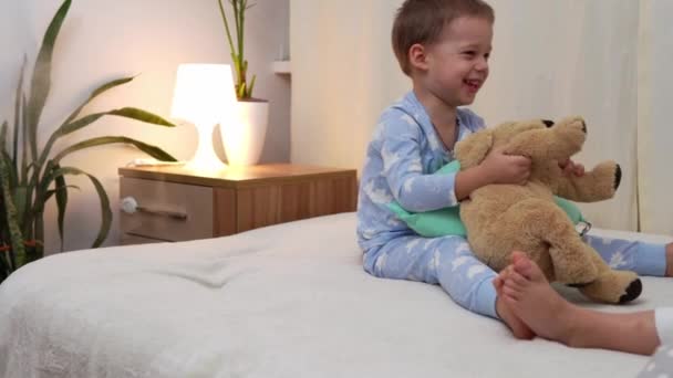 Két mosolygós óvodás gyerek pizsamában játszik Teddy Bear-rel az ágyon. Testvérek Kis Ikrek Fiú és Lány Jó Szórakozást. Boldog gyerekek karanténban otthon. Barátság, család, oktatási koncepció