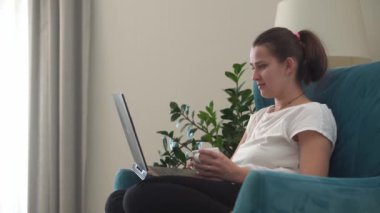 Oturma odasında dizüstü bilgisayarla konuşan otantik bir genç kadın. Kullanarak Yazma Araması. Bilgisayarda çalışan mutlu bayan rahat rahat koltukta interneti tarıyor. Buisenes, Eğitim, Teknoloji Konsepti