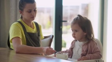 Yakından anne, beyaz giyen, yüksek sandalyedeki bebeği besliyor, çocuk kendini yemeye çalışıyor, yemek lekeli mutlu çocuk, küçük kız kaşıkla yulaf lapası yiyor. Mutfakta kaliteli kahvaltı.