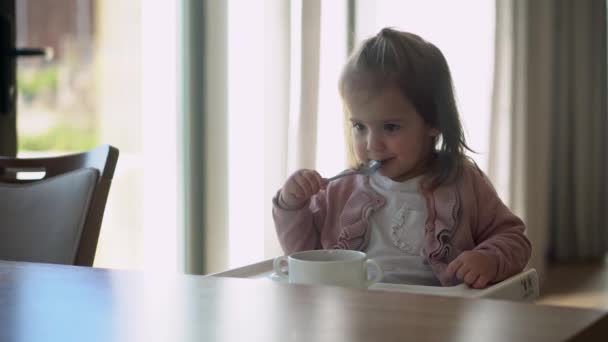 Zbliżenie małego dziecka w białym karmieniu wysokiego krzesełka, dziecko próbuje zjeść siebie, szczęśliwe dziecko z poplamioną jedzeniem twarzą, mała dziewczynka jedząca owsiankę łyżką. Wysokiej jakości karmienie śniadanie w — Wideo stockowe