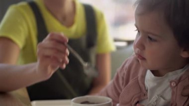 Yakından anne, beyaz giyen, yüksek sandalyedeki bebeği besliyor, çocuk kendini yemeye çalışıyor, yemek lekeli mutlu çocuk, küçük kız kaşıkla yulaf lapası yiyor. Mutfakta kaliteli kahvaltı.
