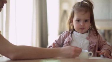 Yakından anne, beyaz giyen, yüksek sandalyedeki bebeği besliyor, çocuk kendini yemeye çalışıyor, yemek lekeli mutlu çocuk, küçük kız kaşıkla yulaf lapası yiyor. Mutfakta kaliteli kahvaltı.