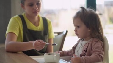 Yakından anne, beyaz giyen, yüksek sandalyedeki bebeği besliyor, çocuk kendini yemeye çalışıyor, yemek lekeli mutlu çocuk, küçük kız kaşıkla yulaf lapası yiyor. Mutfakta kaliteli kahvaltı.