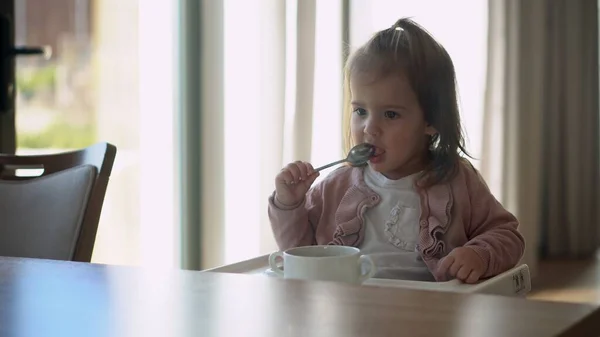 Closeup of young baby in white feeding high chair, kid is trying to eat himself, happy child with food stained face, little Girl eats porridge with spoon. High quality feeding up breakfast in kitchen — Stock Photo, Image