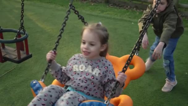 Plano médio de engraçadas meninas pré-escolares em balançar no balanço antes do pôr do sol. Irmãs brincando no parque infantil em Park. Entretenimento para crianças, Infância, Natureza, Conceito de família feliz — Vídeo de Stock