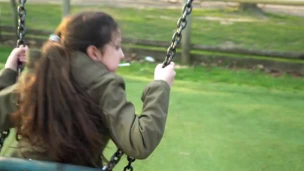 Plano médio de engraçado pouco pré-escolar gordinha menina na jaqueta balançando no balanço antes do pôr do sol. Teen Playing On Playground in Park (em inglês). Entretenimento para crianças, Infância, Natureza, Conceito de família feliz — Vídeo de Stock