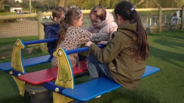小さな幸せなグループ異なる年齢の子供たちはメリーゴーランドで楽しい回転をしています。笑顔外で遊んでいる兄弟の友人。感情的な子供たちは公園の遊び場でカルーセルに乗っています幸せな家族、子供時代 — ストック動画
