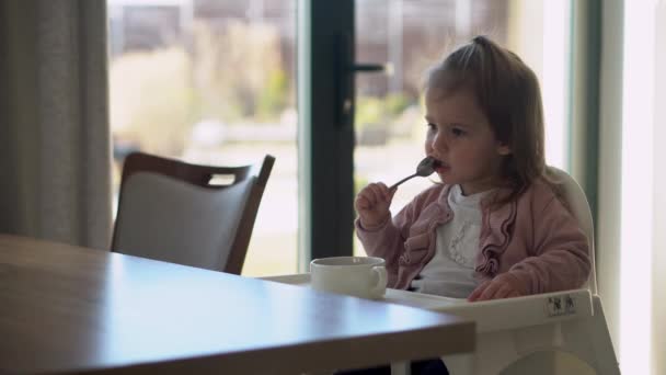 Närbild av ung bebis i vit matning barnstol, ungen försöker äta sig själv, lycklig barn med mat fläckig ansikte, liten flicka äter gröt med sked. Hög kvalitet utfodring frukost i — Stockvideo