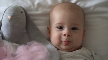 Yakın plan Mutlu neşeli çocuk 6 aylık. Yeni doğan çocuk, beyaz yumuşak yatakta banyo sonrası kameraya bakıyor. Bebek oyuncakla yatmadan önce uyandı. Çocukluk, annelik, aile, bebek konsepti