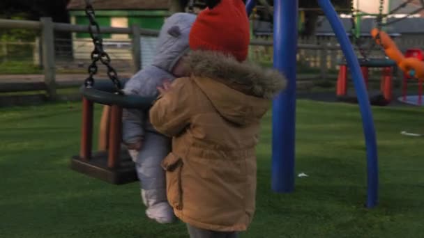 Grappig klein kleuterschooltje mollig meisje ouder zuster rijdt jongere pasgeboren broertje jongen op schommel. Baby spelen op Playground. Kinderanimatie, Kindertijd, Kinderontwikkeling, Happy Family Concept — Stockvideo