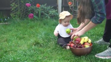 Panama şapkalı küçük bir çocuk büyük bir kâse taze meyveyle çıplak ayakla çimlerin üzerinde oturuyor. Bebek yürümeye başlayan çocuk ısırır elmaları, muz bahçesini, bahçeyi, sağlıklı yiyecekleri yer.