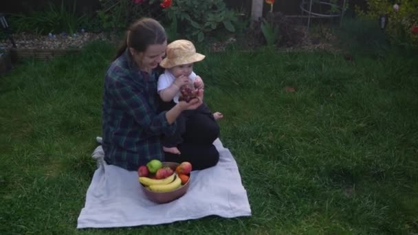 Счастливая молодая веселая мать держит ребенка едят фрукты на зеленой траве. Мама прелестный младенец играет на свежем воздухе с любовью в саду. "Малыш с родителями". Семья, природа, экология — стоковое видео