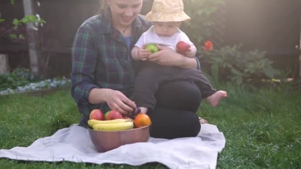 Szczęśliwa młoda radosna matka trzymająca dziecko jedzące owoce na zielonej trawie. Mama urocze dziecko bawiące się na dworze z miłością w ogrodzie. Mały dzieciak z rodzicami. Pojęcie rodziny, przyrody, ekologii — Wideo stockowe