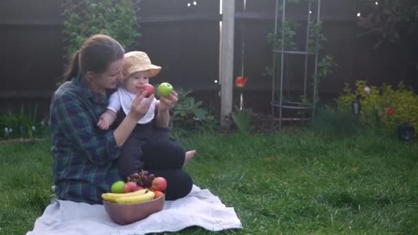 Boldog fiatal, vidám anya tartja baba eszik gyümölcsöt a zöld fű. Anya imádnivaló csecsemő gyermek játszik a szabadban szeretettel a kertben. Kisgyerek a szülőkkel. Család, természet, ökológia koncepció — Stock videók