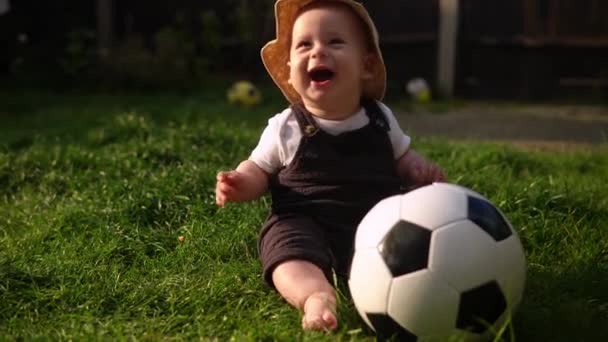 Happy Baby Sitting With Soccer Black Classic Ball On Green Grass Приваблива дитина бавиться на подвір'ї. Малі діти з батьками. Футбол, чемпіонат, спортивна концепція — стокове відео