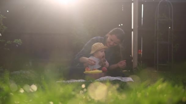 Счастливая молодая веселая мать держит ребенка едят фрукты на зеленой траве. Мама прелестный младенец играет на свежем воздухе с любовью в саду. "Малыш с родителями". Семья, природа, экология — стоковое видео