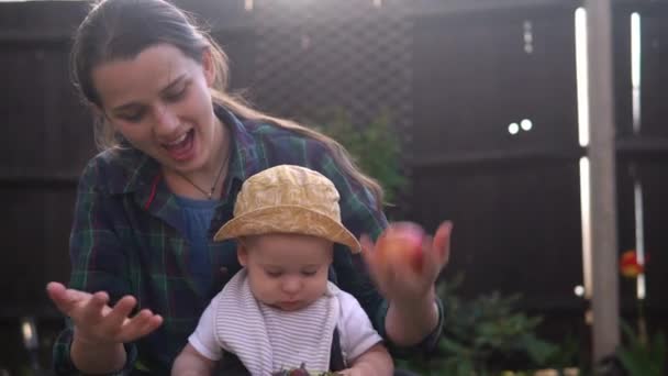 Šťastná mladá veselá matka drží dítě jíst ovoce na zelené trávě. Máma rozkošné dítě hrát venku s láskou v zahradě. Malý kluk s rodiči. Rodina, příroda, koncepce ekologie — Stock video
