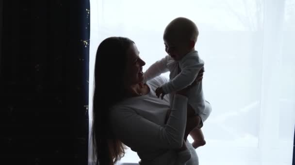 Silhouette amorevole tenera caucasico giovane mamma holding adorabile carino bambino ragazzo figlio abbracciare baciare piccolo bambino. felice affettuoso lungo dai capelli madre coccole con infantile bambina figlia a casa. — Video Stock