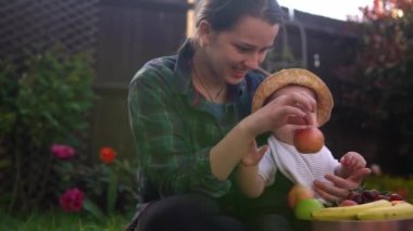 Mutlu Genç Neşeli Anne Yeşil Çimlerde Bebek Yiyen Meyveler Tutuyor. Sevimli Anne Bebek Arka Bahçe Bahçesinde Dışarıda Sevgiyle Oynuyor. Ailesi olan küçük çocuk. Aile, Doğa, Ekoloji Kavramı