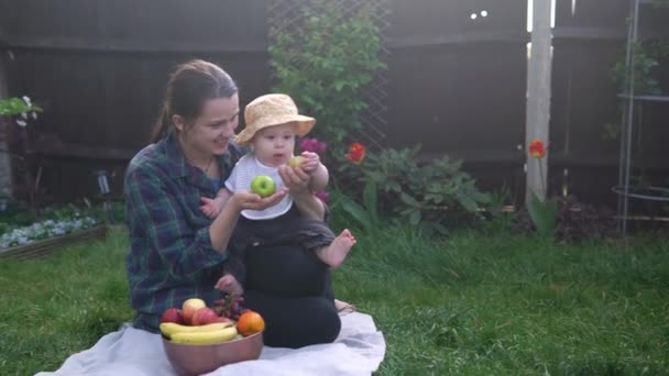 快乐的年轻快乐的母亲抱着孩子在绿草上吃水果。妈妈可爱的婴儿在后院花园里玩着爱的户外游戏。和父母在一起的小孩家庭、自然、生态概念 — 图库视频影像