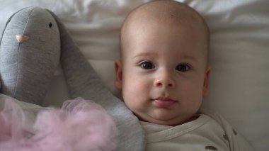 Yakın plan Mutlu neşeli çocuk 6 aylık. Yeni doğan çocuk, beyaz yumuşak yatakta banyo sonrası kameraya bakıyor. Bebek oyuncakla yatmadan önce uyandı. Çocukluk, annelik, aile, bebek konsepti