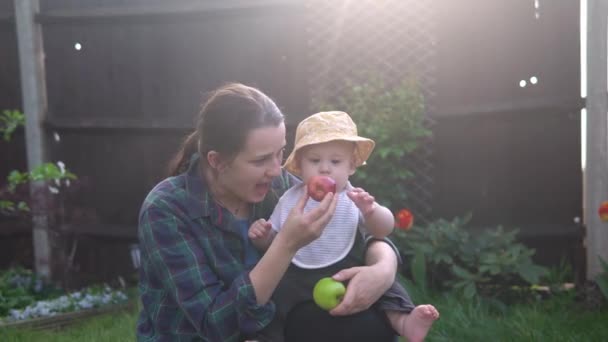 Šťastná mladá veselá matka drží dítě jíst ovoce na zelené trávě. Máma rozkošné dítě hrát venku s láskou v zahradě. Malý kluk s rodiči. Rodina, příroda, koncepce ekologie — Stock video